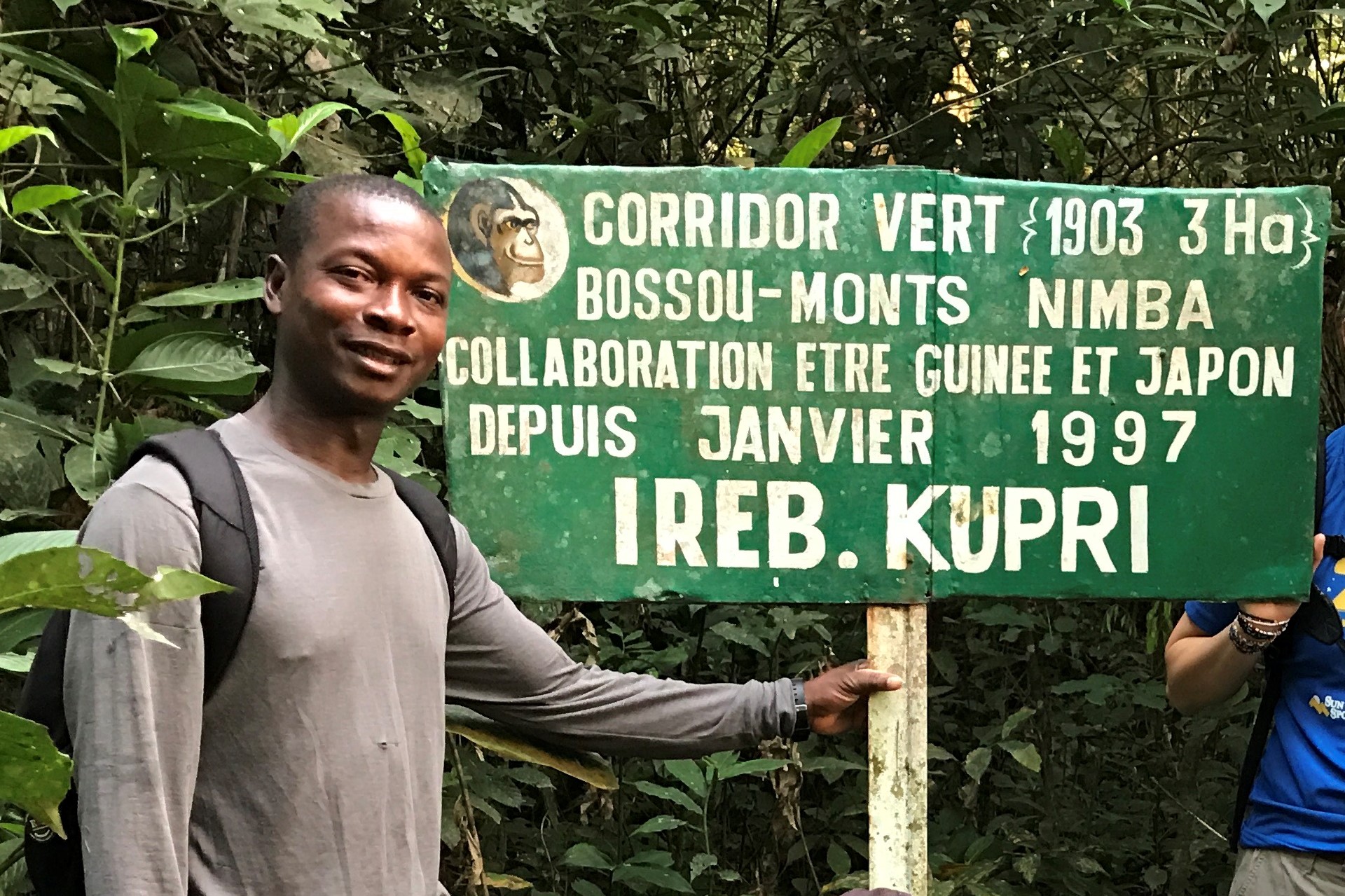 Bossou field assistant Henry Camara stood in front of a green sign that reads "CORRIDOR VERT {1903 3 Ha} BOSSOU-MONTS NIMBA COLLABORATION ETRE GUINEE ET JAPON DEPUIS JANVIER 1997 IREB. KUPRI"