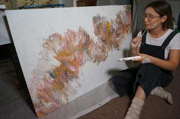 Megan Beardmore-Herd sat in front of a painted board, brush in hand, paint on face, contemplating the next brush stroke